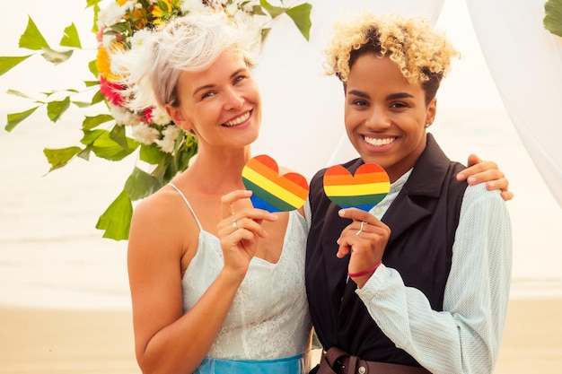 Sposo femminile afroamericano in abito nero e sposa caucasica in abito in cerimonia sulla spiaggia tropicale sotto l'arco di nozze