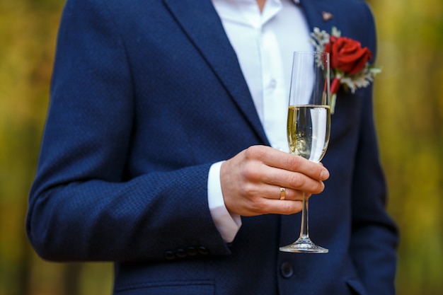 Sposo in abito blu con boutonniere rosso tenere tra le mani bicchieri da vino con champagne