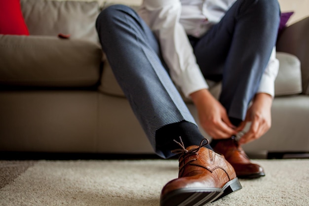 Sposo in pantaloni blu e camicia bianca seduto sul divano ad allacciarsi i lacci delle scarpe marroni