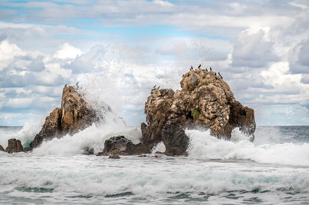 Spruzzata dell'acqua dell'oceano sulla spiaggia rocciosa con bel cielo e nuvole. Onda del mare che spruzza sulla pietra in riva al mare in inverno. La linea delle ciglia delle onde del mare impattano la roccia sulla spiaggia