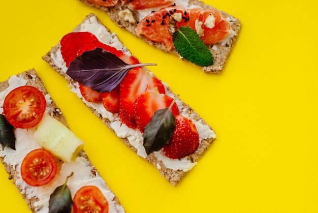 Spuntino sano con pane croccante, crema di formaggio, fragola, pompelmo, pomodoro e cetriolo su uno sfondo giallo