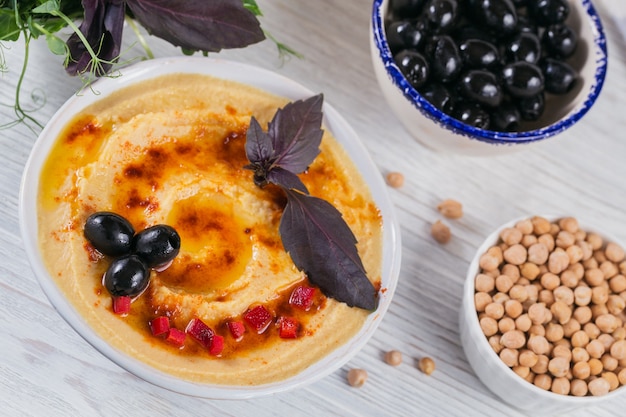 Spuntino sano da pane croccante con hummus, olio d'oliva, olive nere e paprika su una superficie di legno bianca.