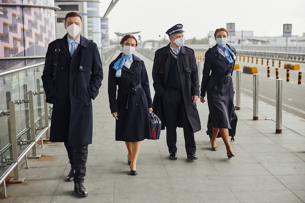 Squadra dell'aereo che cammina con i bagagli sul marciapiede vicino all'aeroporto. Donne e uomini indossano uniformi e maschere mediche. Pilota, assistente di volo e hostess. Lavoro di squadra. Aviazione civile. Concetto di viaggio aereo
