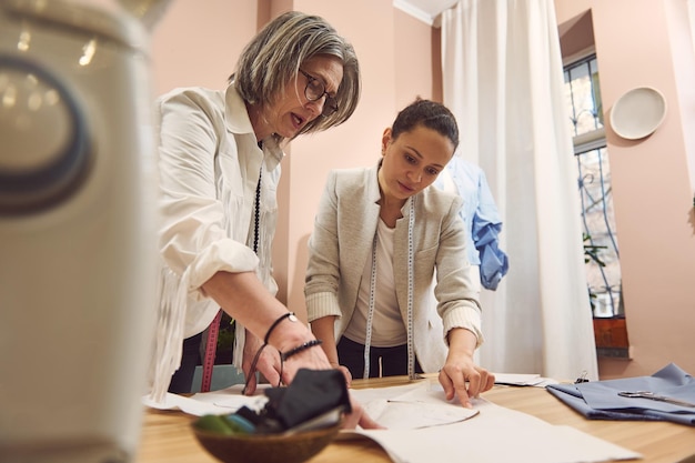 Squadra di due stiliste multietniche che lavorano alla raccolta di modelli di cucito per creare una nuova collezione di abiti moderni alla moda in un atelier di sartoria Sarte che discutono di modelli