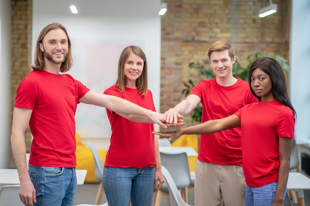 Squadra. Gruppo di giovani sorridenti in magliette rosse e jeans in piedi allungando la mano insieme in avanti all'interno