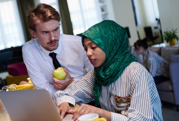 squadra internazionale di affari multiculturali. uomo che mangia mela donna musulmana africana che indossa un hijab verde che beve tè mentre lavora insieme su un computer portatile nell'area relax al moderno piano aperto startu