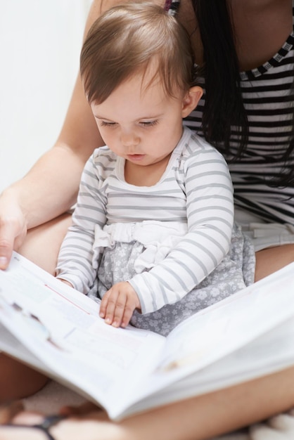 Sta già imparando Inquadratura di una bambina carina seduta seduta sulle ginocchia di sua madre che guarda attraverso un libro illustrato