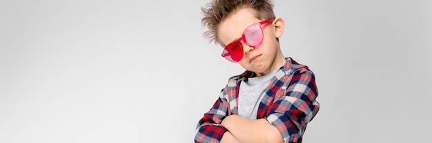 Sta un bel ragazzo in camicia a quadri, camicia grigia e jeans. Un ragazzo in occhiali da sole rossi. Il ragazzo incrociò le braccia sul petto.