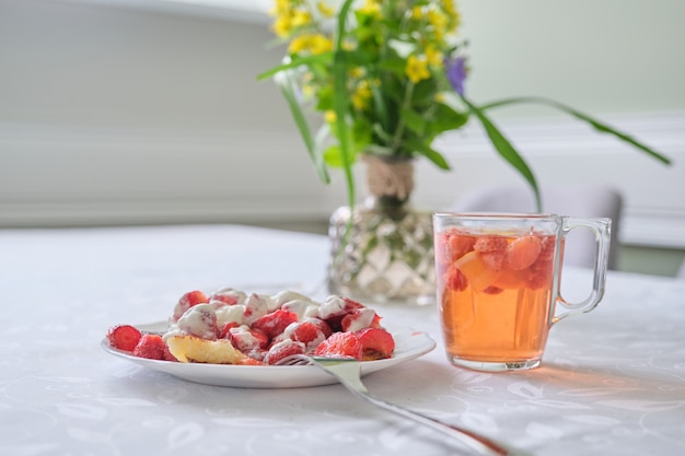 Stagione delle fragole, cibi e bevande con frutti di bosco. Cibo sul tavolo, frittelle di cagliata con fragole e panna acida. Tè al limone e frutti di bosco