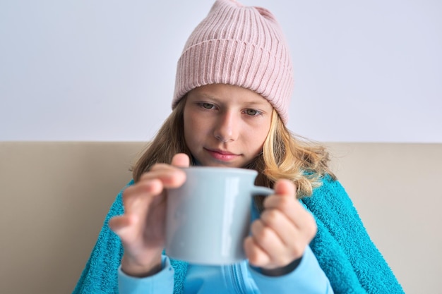 Stagione fredda autunno inverno, bambina seduta a casa con una tazza di tè caldo, riscaldamento in berretto lavorato a maglia sotto una coperta calda