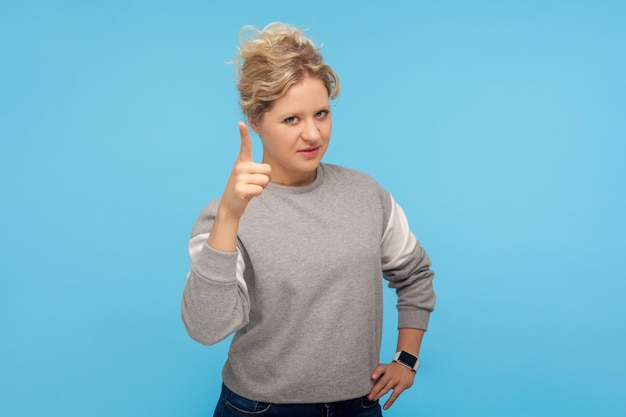 Stai attento. Donna arrabbiata seria con capelli ricci corti in felpa che avverte con il dito alzato, gesto ammonente, incolpandoti per errore, rimproverando. tiro in studio indoor isolato su sfondo blu