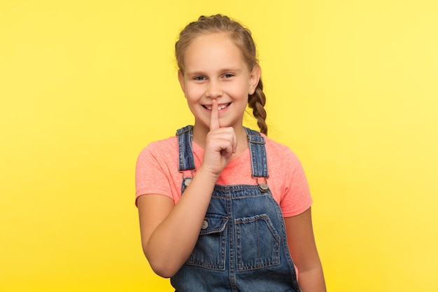 Stai zitto, è segreto. Ritratto di bambina positiva con treccia in tuta di jeans che fa un gesto di silenzio con il dito sulle labbra, mistero del bambino. tiro in studio indoor isolato su sfondo giallo