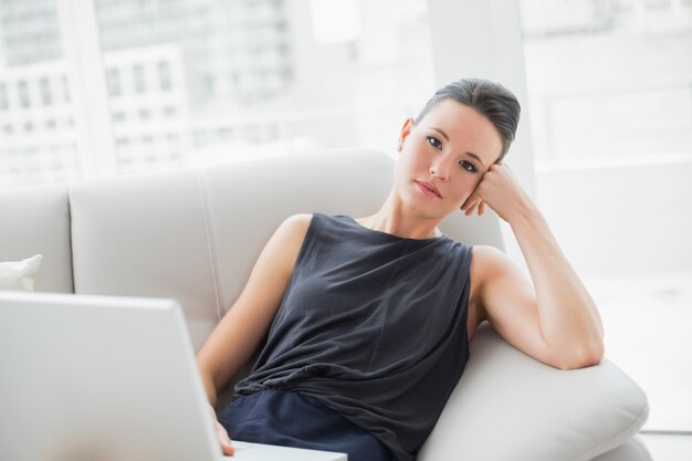 Stanca bella donna ben vestita con il portatile seduto sul divano