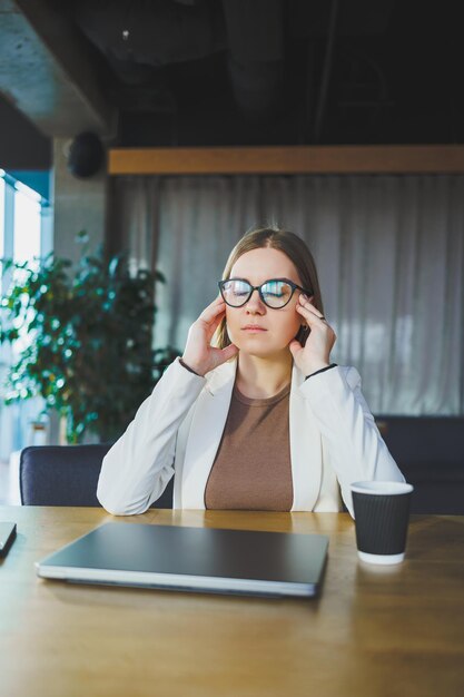 Stanca giovane donna d'affari seduta in ufficio a un tavolo di legno dietro un computer portatile Un giovane manager con gli occhiali è stanco del lavoro