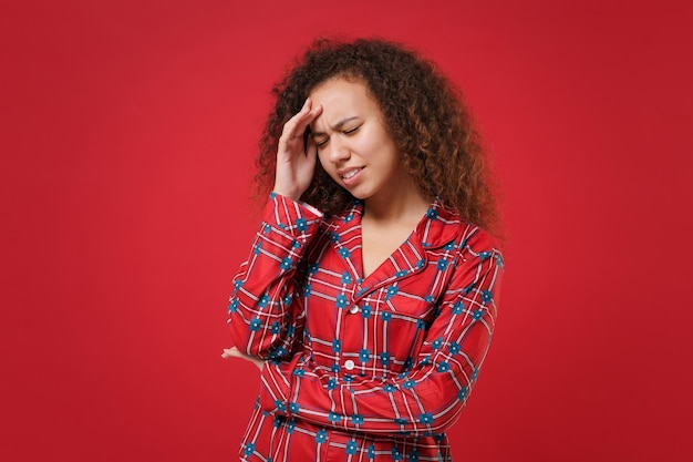 Stanca ragazza afroamericana in pigiama homewear che riposa a casa isolata su sfondo rosso. Rilassatevi il concetto di stile di vita di buon umore. Mock up spazio di copia. Tenere gli occhi chiusi, mettere la mano sulla testa, avere mal di testa.