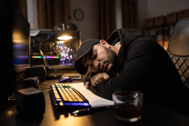 Stanco assonnato oberato di lavoro con berretto e felpa lavora di notte in una stanza buia dorme sulla scrivania