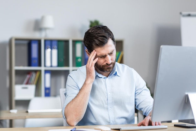 Stanco e infastidito giovane impiegato manager che tiene la testa sentendo dolore vertigini Seduto alla scrivania di un computer in ufficio