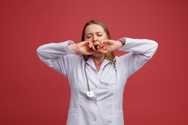 Stanco giovane bionda femmina medico indossa veste medica e stetoscopio intorno al collo mantenendo le mani sulla bocca che sbadiglia con gli occhi chiusi