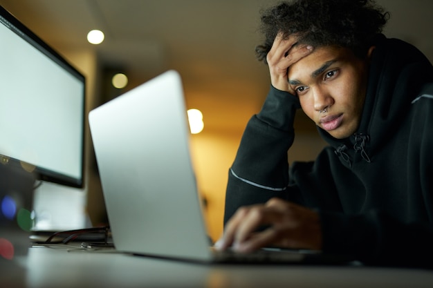 Stanco giovane ragazzo che studia o lavora al computer portatile seduto in una stanza buia a casa a tarda notte