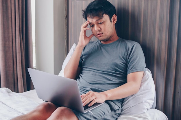 Stanco stressato giovane asiatico che si sente assonnato e stanco mentre usa il laptop sul letto in camera da letto Concetto di duro lavoro