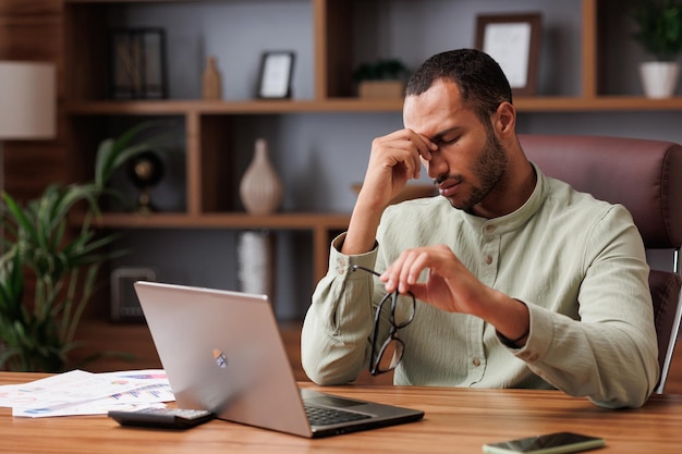 Stanco uomo d'affari afroamericano dopo aver esaurito le scartoffie togliendosi gli occhiali e sentendo affaticamento degli occhi Mal di testa cattiva visione affaticamento cronico e affaticamento degli occhi concetto Burnout e concetto di superlavoro