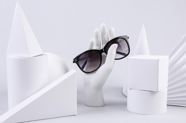Stand mano che tiene occhiali da sole e forme geometriche su sfondo bianco Concept art Minimalismo