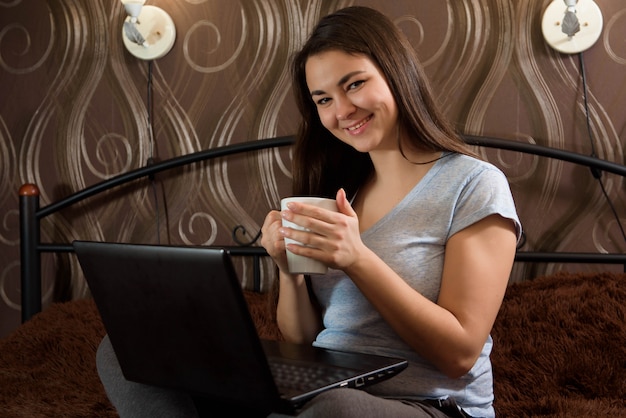 Stare a casa! Lavoro a casa. Bella donna casuale che lavora ad un computer portatile che si siede sul letto.