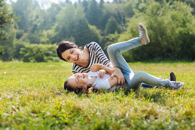 Stato d'animo gioioso. Felice donna affascinante scherzare con la sua piccola figlia mentre giaceva sull'erba nel prato del parco inzuppato di sole