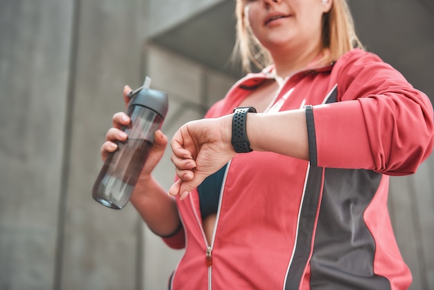 È stato un ottimo allenamento. Giovane donna plus size in abbigliamento sportivo che controlla il contapassi e tiene in mano una bottiglia d'acqua stando in piedi all'aperto. Perdere peso. Concetto di sport