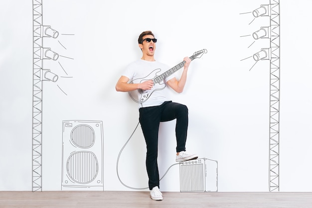 Stella del rock! Giovane bell'uomo con gli occhiali da sole che suona la chitarra disegnata e tiene la bocca aperta mentre si trova su uno sfondo bianco con l'illustrazione della colonna stereo e della luce del palcoscenico stage