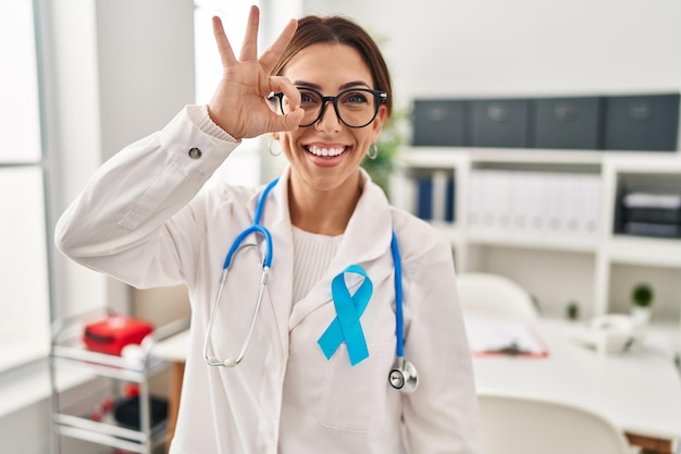 Stetoscopio da portare della giovane donna di medico castana alla clinica che fa gesto giusto con l'occhio sorridente della mano che guarda attraverso le dita con la faccia felice