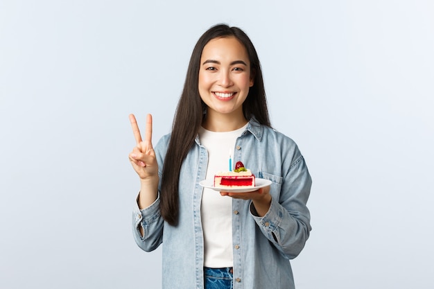 Stile di vita a distanza sociale, pandemia di covid-19, celebrazione delle vacanze durante il concetto di coronavirus. Felice compleanno ragazza asiatica sorridente che tiene la torta di compleanno e mostra il segno di pace