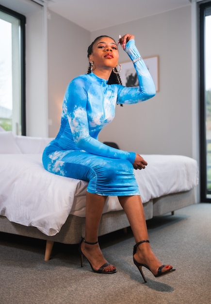 Stile di vita alla moda di una ragazza etnica nera con i capelli raccolti e un vestito blu, seduto su un letto d'albergo