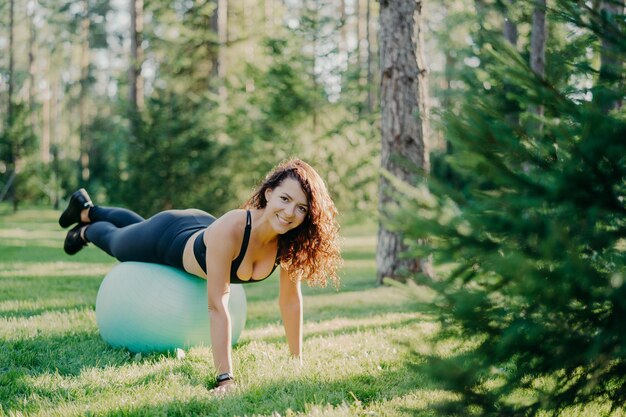 Stile di vita attivo fitness e concetto di cura del corpo Fit felice giovane donna bruna indossa esercizi di abbigliamento sportivo con palla fitness pone nella foresta su erba verde Sport pilates esercizio