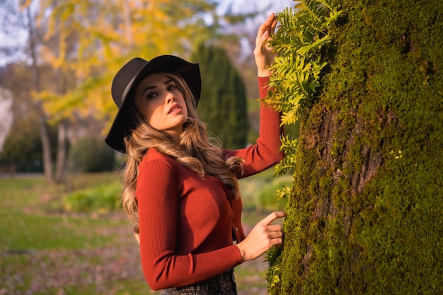 Stile di vita bionda ragazza caucasica in un maglione rosso e cappello nero godersi la natura in un parco con alberi ritratto di giovane donna che guarda il bellissimo albero con muschio in autunno
