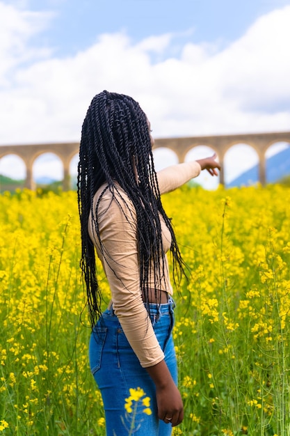 Stile di vita che punta al ritratto del ponte di una ragazza etnica nera con trecce viaggiatore in un campo di fiori gialli