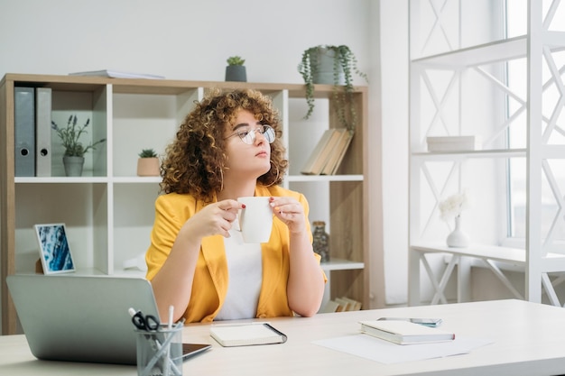 Stile di vita da freelance Lavorare da casa Pausa caffè Donna d'affari in sovrappeso rilassata alla scrivania con il laptop che si gode il tempo libero guardando la finestra all'interno del posto di lavoro leggero