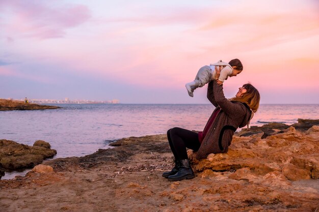 Stile di vita di una famiglia sulla spiaggia un bambino che si diverte e lo solleva con sua madre seduta in riva al mare al tramonto