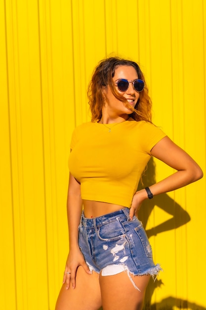 Stile di vita, ragazza bionda caucasica in magliette gialle e jeans corti su una parete gialla. Posa alla moda con un sorriso