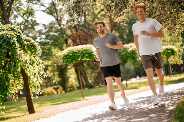 Stile di vita. Ragazzi energici e attraenti in pantaloncini e maglietta che corrono di buon umore nel parco verde la mattina d'estate