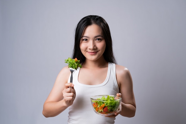 Stile di vita sano con il concetto di cibo pulito. Donna asiatica felice che mangia insalata isolata sopra il muro bianco.