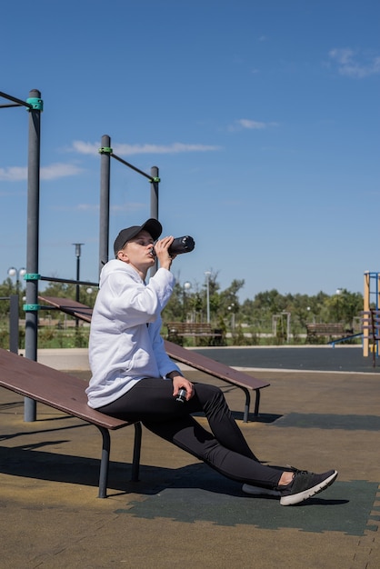 Stile di vita sano e attivo. Sport e fitness. Giovane donna sportiva in abiti sportivi che beve acqua dal thermos sul campo sportivo