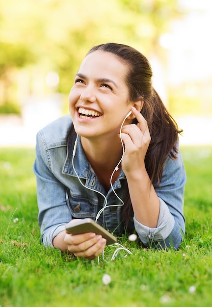 stile di vita, vacanze estive, tecnologia, tempo libero e concetto di persone - ragazza ridente con smartphone e auricolari sdraiato sull'erba