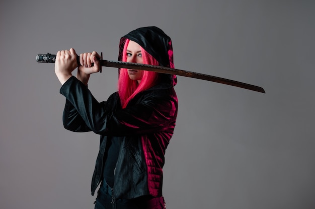 Stile techno cyber punk, ninja futuristico, giovane donna con capelli rosa in abiti neri con una katana, foto in studio su grigio