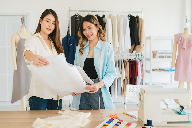 Stilista o sarto femminile asiatico con assistenza nella preparazione del modello di tessuto di carta tagliata Sarta che lavora e progetta nuove collezioni di moda per i clienti nel laboratorio di sartoria dell'atelier