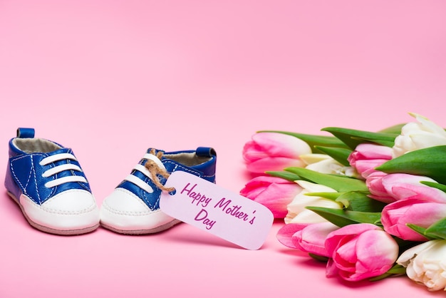 Stivaletti per bambini con scritta Happy Mothers Day sull'etichetta di carta vicino al bouquet di tulipani sulla superficie rosa