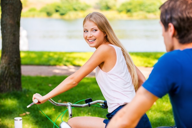 sto vincendo! Bella giovane donna sorridente in sella alla sua bicicletta e guardando sopra la spalla mentre il suo ragazzo cavalca dietro di lei