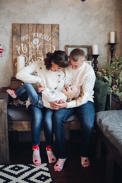 Stock photo ritratto di genitori felici in maglioni bianchi solletico la loro piccola figlia in ginocchio. La figlia sta ridendo allegramente. Decorazioni natalizie.