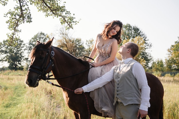 Storia d'amore e matrimonio vicino al fiume in un campo al tramonto con cavallo marrone. La sposa in abito arioso è il colore della rosa polverosa. Abito beige con scintillii. la sposa e lo sposo abbracciano e baciano.