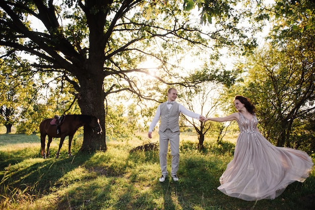 Storia d'amore e matrimonio vicino al fiume in un campo al tramonto con cavallo marrone. La sposa in abito arioso è il colore della rosa polverosa. Abito beige con scintillii.